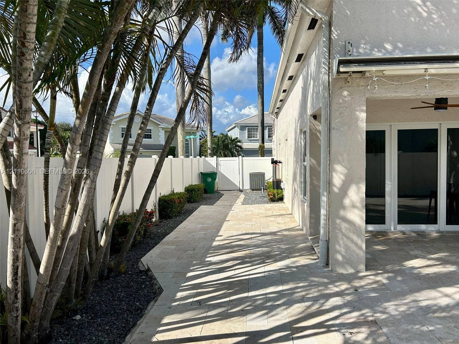 Covered patio featuring stone-paver flooring, white vinyl perimeter fencing, and established palm trees