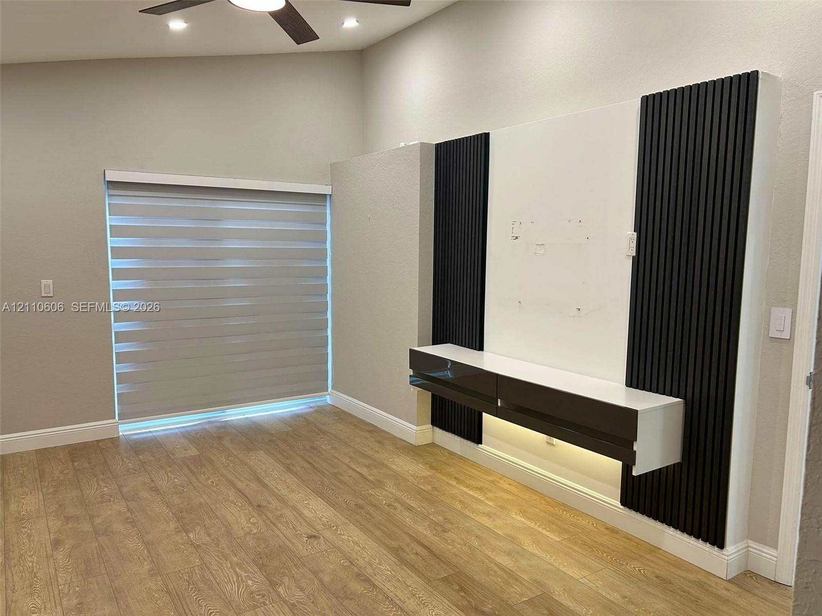 Vaulted ceiling featuring a ceiling fan, wood-finish flooring, and a custom built-in media console with black vertical accent paneling and integrated under-cabinet lighting