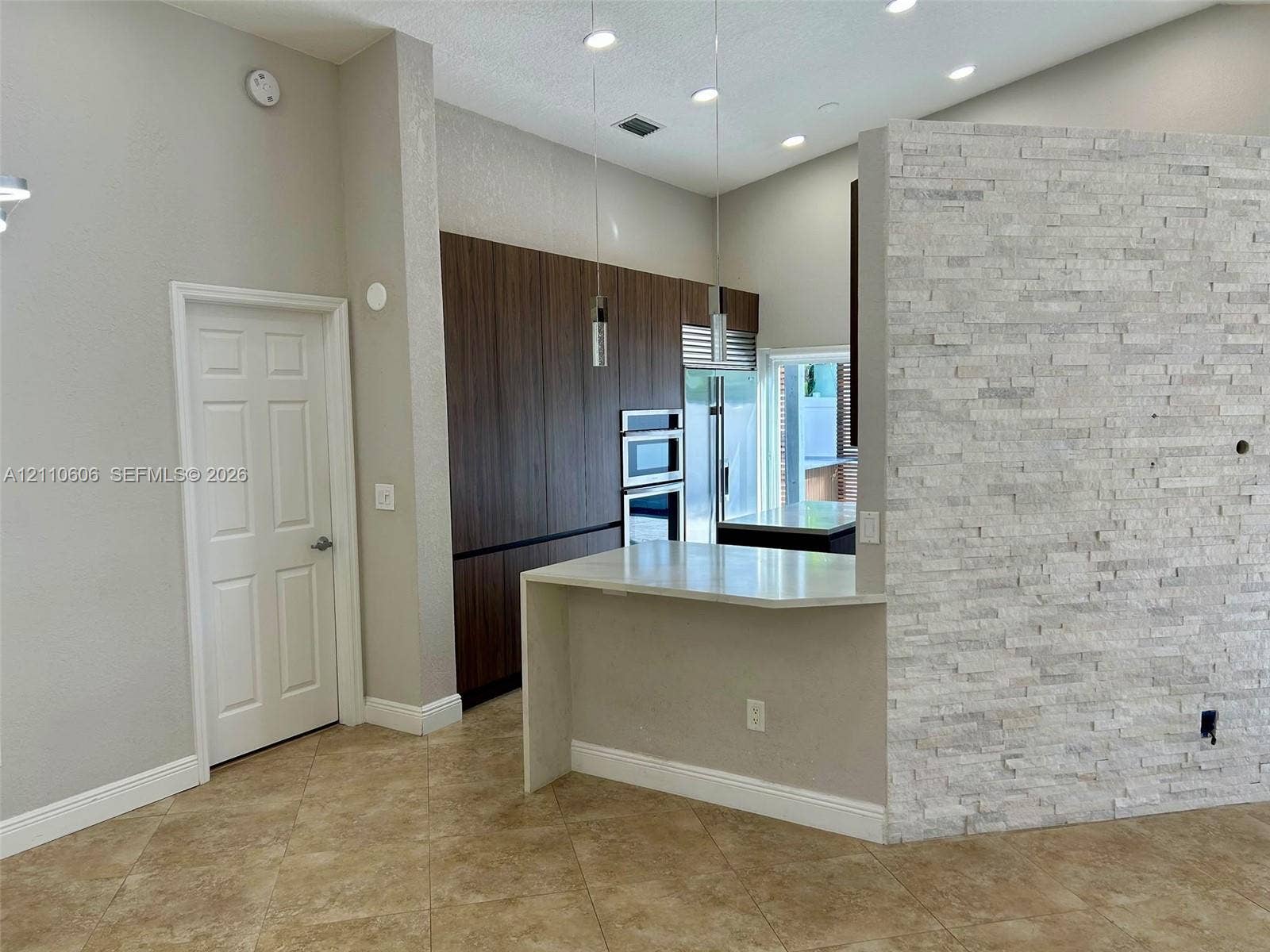 Kitchen featuring flat-panel wood-finish cabinetry, built-in double ovens, a peninsula with a waterfall-edge countertop, and a decorative stone-veneer accent wall