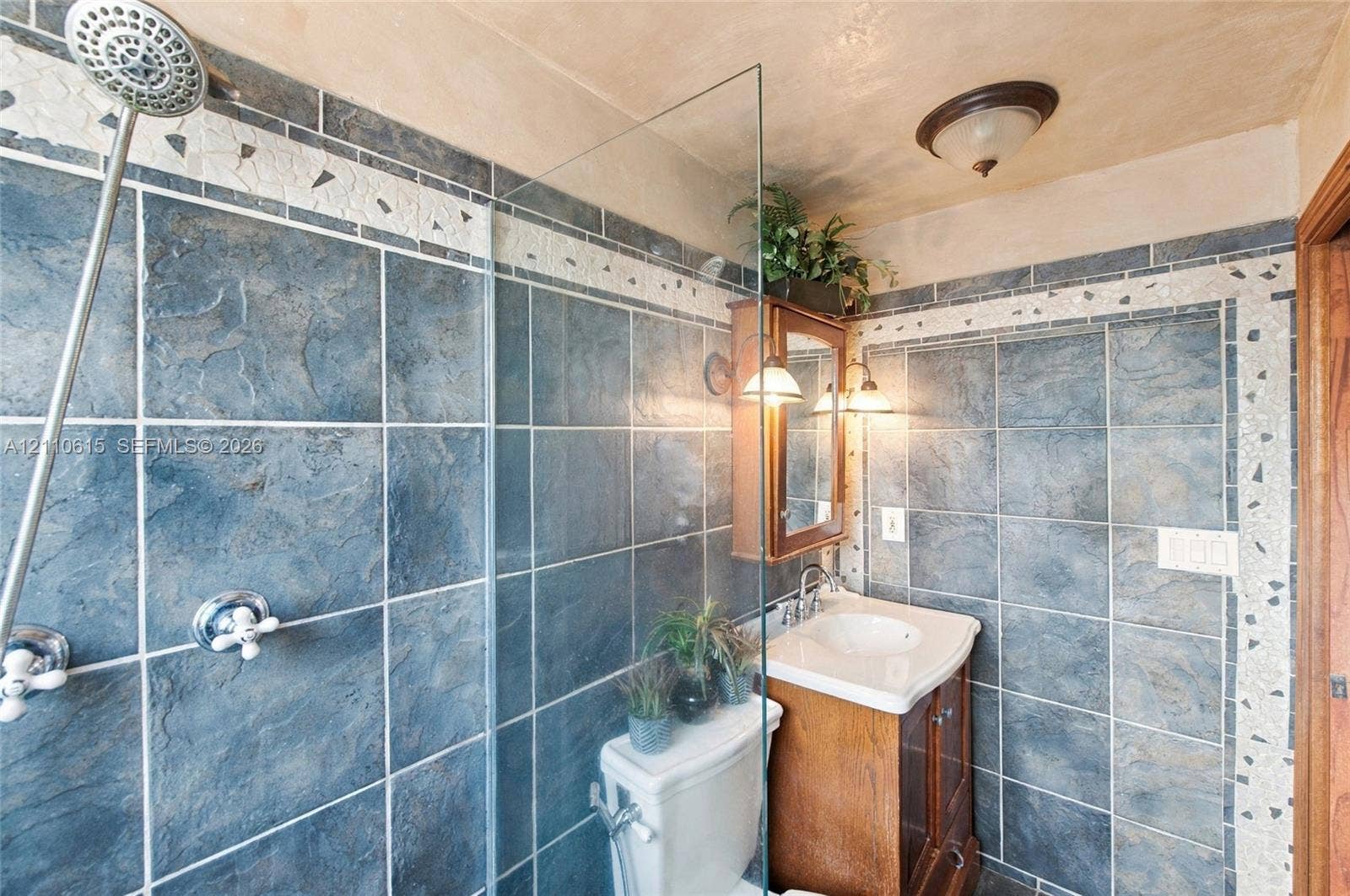 Walk-in shower with floor-to-ceiling stone-finish tile, glass partition, integrated mosaic tile detailing, pedestal vanity with porcelain sink, and wall-mounted light fixtures