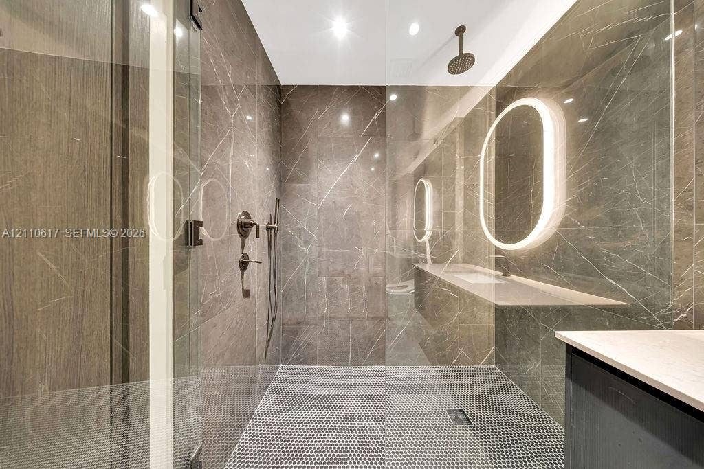 Walk-in shower featuring floor-to-ceiling stone-finish tile, a rainfall showerhead, a frameless glass enclosure, a penny-tile floor, and an integrated illuminated oval mirror