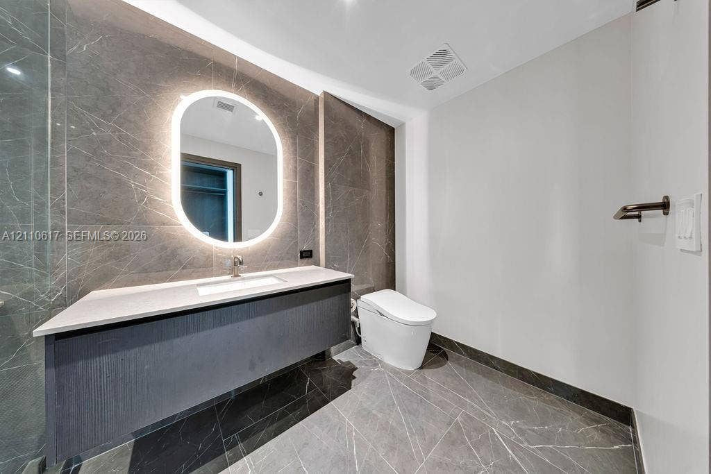 Floating vanity featuring a stone-topped counter and integrated sink, illuminated oval wall mirror, large-format stone tile wall and flooring, and wall-mounted toilet
