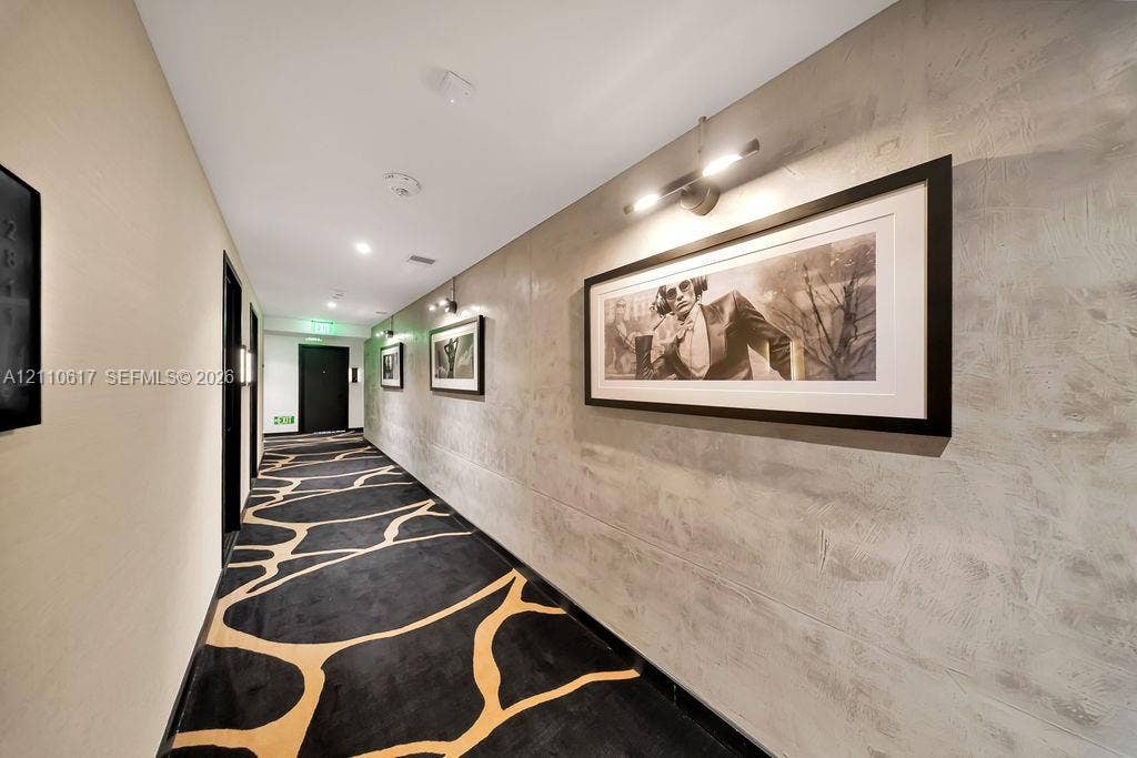 Hallway featuring textured grey wall surfaces, patterned carpeting, and directional wall-mounted lighting