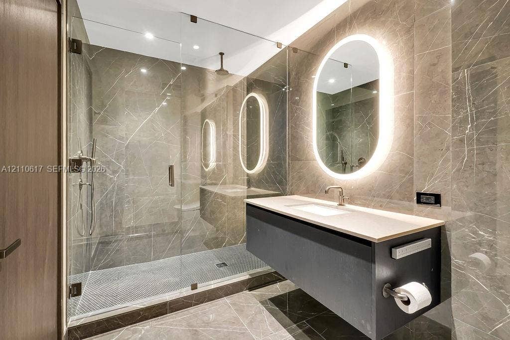 Floating vanity with a light-toned countertop, floor-to-ceiling stone tile wall, walk-in glass-enclosed shower, and oval-shaped backlit mirror