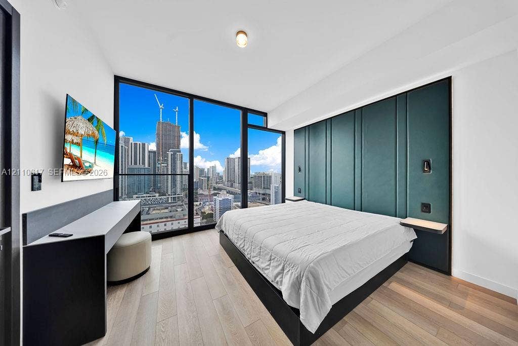 Floor-to-ceiling windows with urban skyline backdrop, custom floor-to-ceiling upholstered teal headboard, built-in desk with integrated shelving, and wide-plank wood-finish flooring