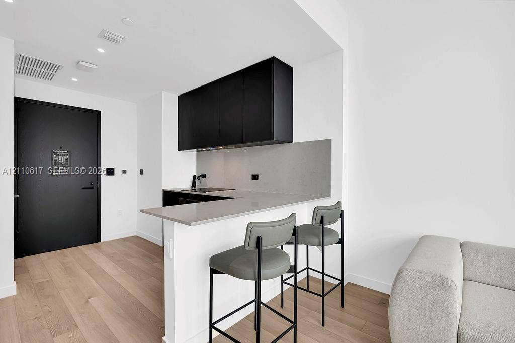 Kitchen area featuring matte black cabinetry, light-toned stone-finish countertops, a matching full-height backsplash, wood-finish flooring, and a black solid-core entry door