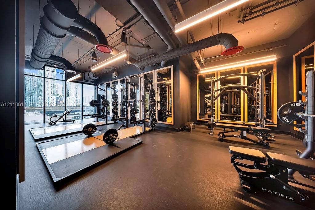 Fitness facility featuring rubber flooring, floor-to-ceiling windows, industrial-style exposed ductwork, wall-mounted mirrors, and integrated linear lighting