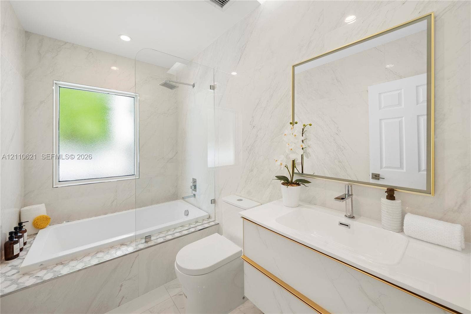 Full-height stone-finish wall tile, white vanity with gold-tone hardware, frameless glass shower enclosure, integrated soaking tub, and gold-framed wall mirror