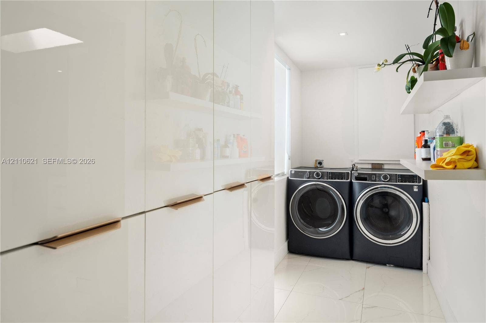 Floor-to-ceiling white high-gloss cabinetry featuring recessed gold-tone pulls, large-format tile flooring, and wall-mounted shelving