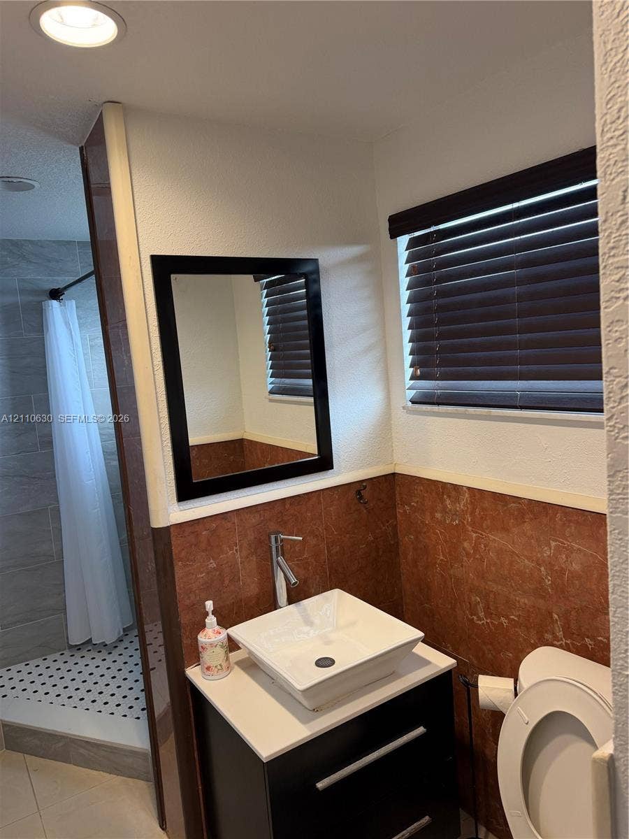 3601 NE 170th Street, #407, North Miami Beach, FL 33160 Modern vanity featuring a square vessel sink with a chrome faucet, topped with a countertop, complemented by a framed mirror, window blinds, and tile wainscoting