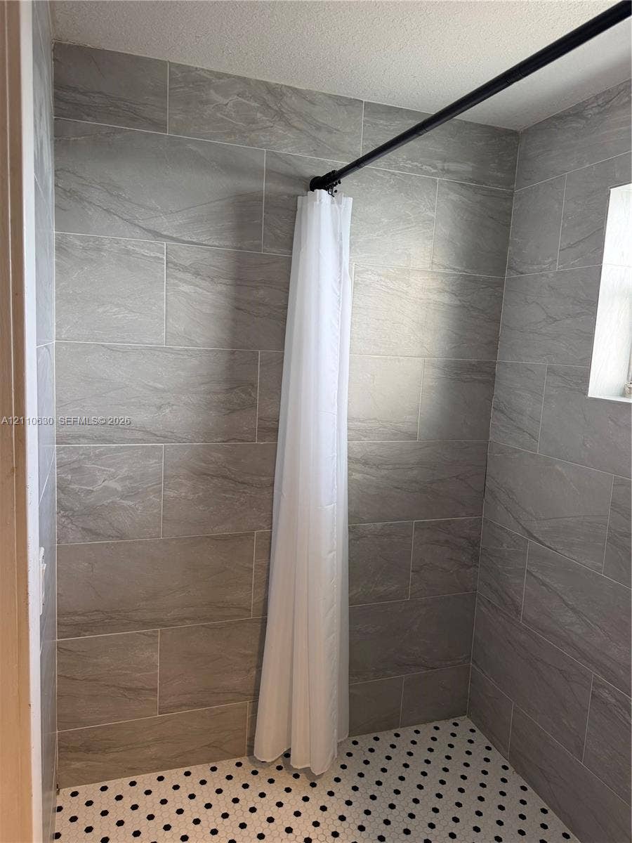 3601 NE 170th Street, #407, North Miami Beach, FL 33160 Walk-in shower enclosure featuring large-format grey tile wall surrounds, hexagonal floor tile with black accent detailing, and a black curtain rod