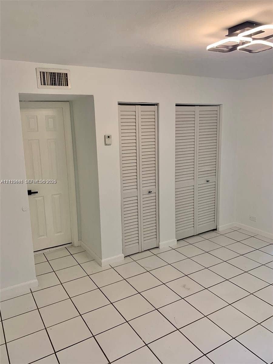 Neutral-toned room featuring large-format square tile flooring, two sets of louvered bifold closet doors, a single-panel interior door with dark hardware, and a modern geometric flush-mount ceiling light