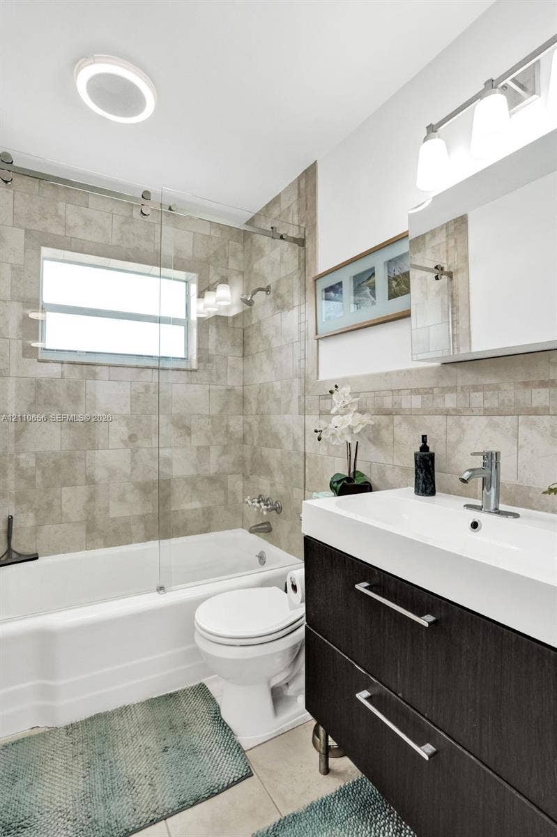 30 NE 48th Court, Oakland Park, FL 33334 Bathroom featuring a modern floating vanity with a rectangular sink, a glass-panel tub enclosure, floor-to-ceiling tile surrounds, and polished chrome fixtures