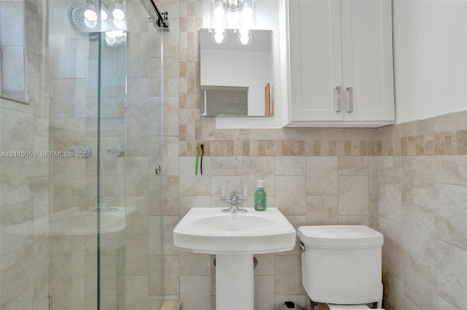 30 NE 48th Court, Oakland Park, FL 33334 Glass-enclosed shower with stone tile surround, pedestal sink with polished chrome hardware, built-in white wall cabinetry, and coordinating stone wall tiling