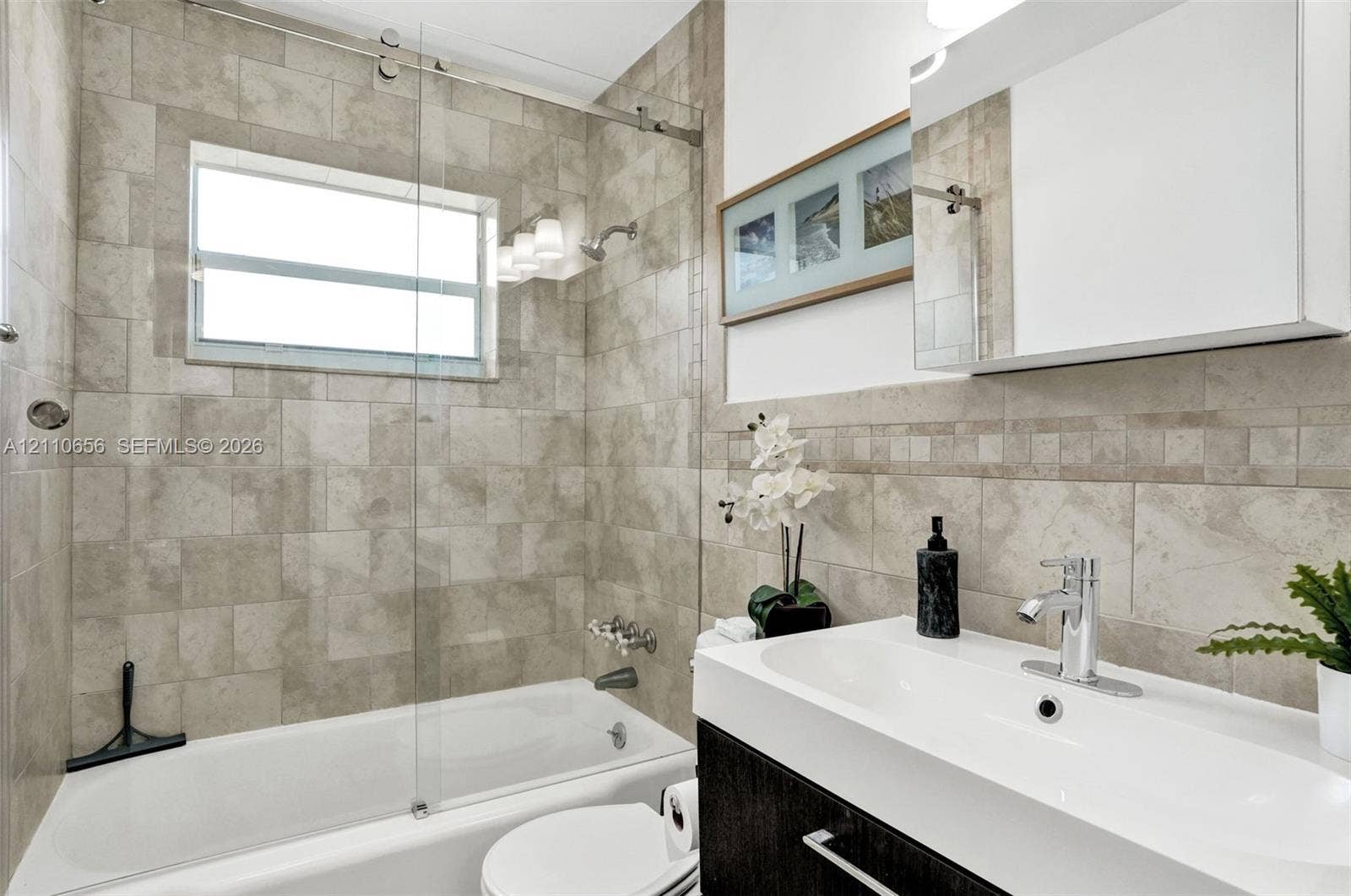 30 NE 48th Court, Oakland Park, FL 33334 Bathroom featuring a glass sliding shower door, neutral wall tile with a mosaic accent border, white undermount sink with chrome fixtures, and dark wood-finish cabinetry