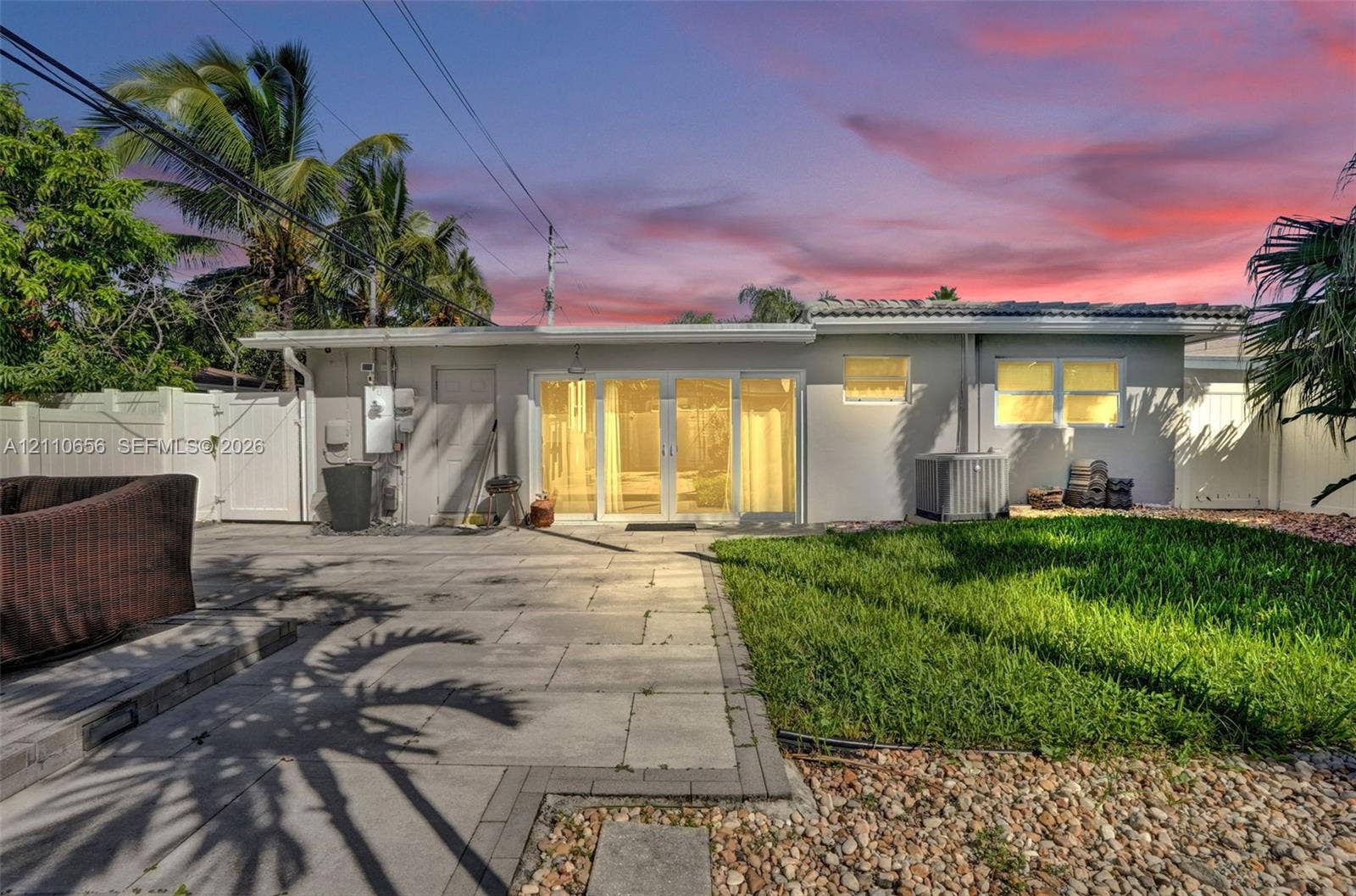 30 NE 48th Court, Oakland Park, FL 33334 Exterior featuring a paved stone patio, multi-panel glass sliding doors, vinyl perimeter fencing, and established palm trees