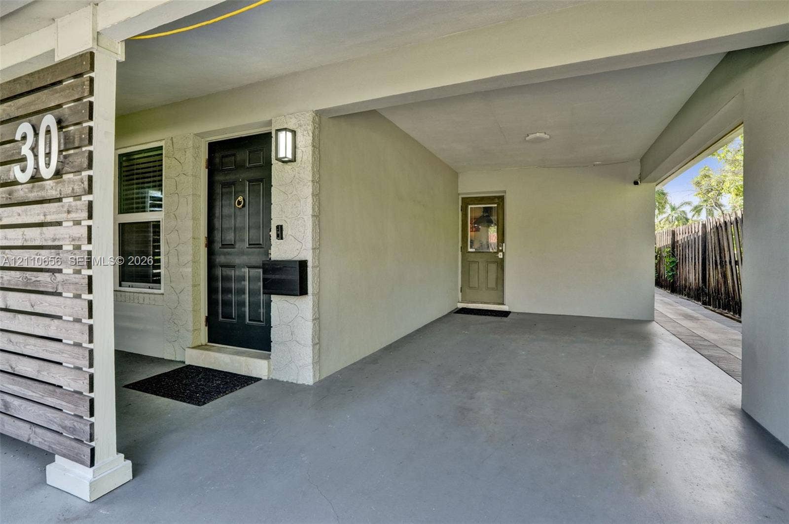 30 NE 48th Court, Oakland Park, FL 33334 Covered carport featuring horizontal wood-slat privacy screening, a dark-toned entry door with a knocker, exterior stone-veneer accents, and a secondary side-access door