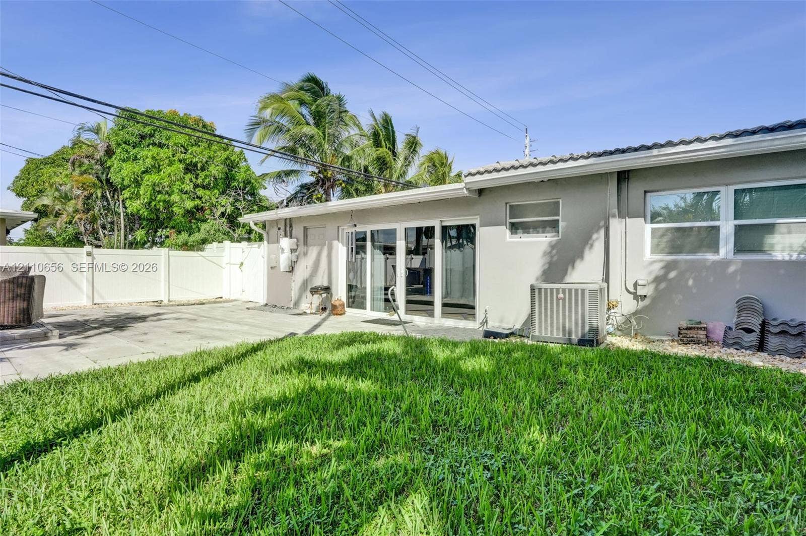 30 NE 48th Court, Oakland Park, FL 33334 Rear exterior featuring a concrete patio, sliding glass doors, a white vinyl privacy fence, and mature palm trees