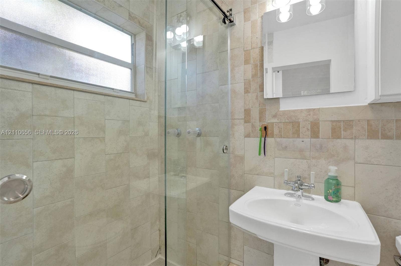 30 NE 48th Court, Oakland Park, FL 33334 Bathroom featuring a frameless glass shower enclosure, neutral-toned stone tile surround, pedestal sink with chrome hardware, and horizontal sliding window
