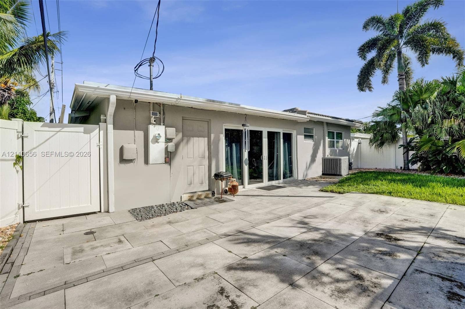 30 NE 48th Court, Oakland Park, FL 33334 Rear exterior featuring a concrete paver patio, sliding glass doors, a solid-panel white privacy gate, and manicured lawn space