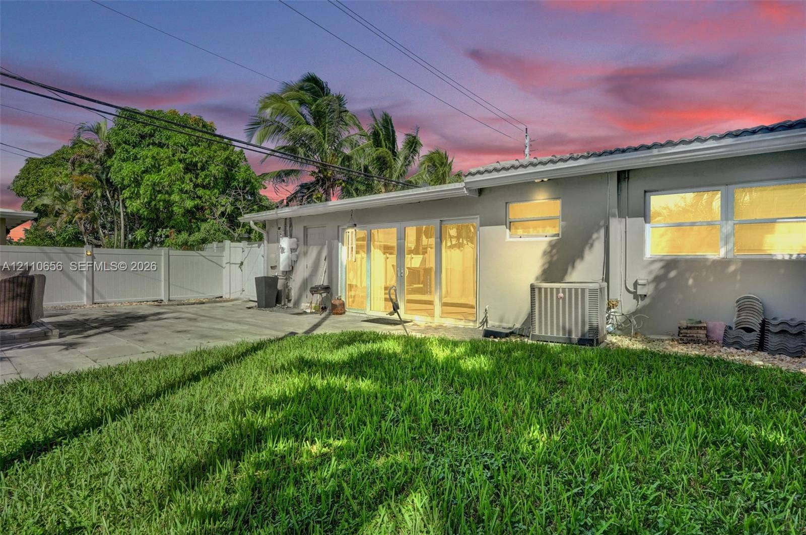 30 NE 48th Court, Oakland Park, FL 33334 Rear exterior featuring a manicured lawn, paved patio area, white vinyl privacy fencing, sliding glass doors, and mature palm trees