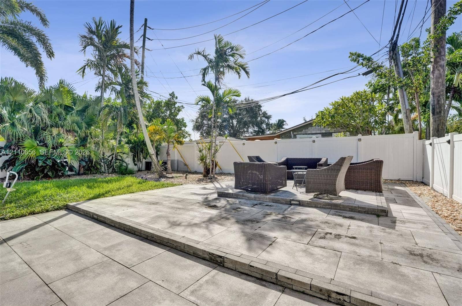 30 NE 48th Court, Oakland Park, FL 33334 Backyard featuring a tiered stone paver patio, white vinyl privacy fencing, mature palm trees, and established landscaping