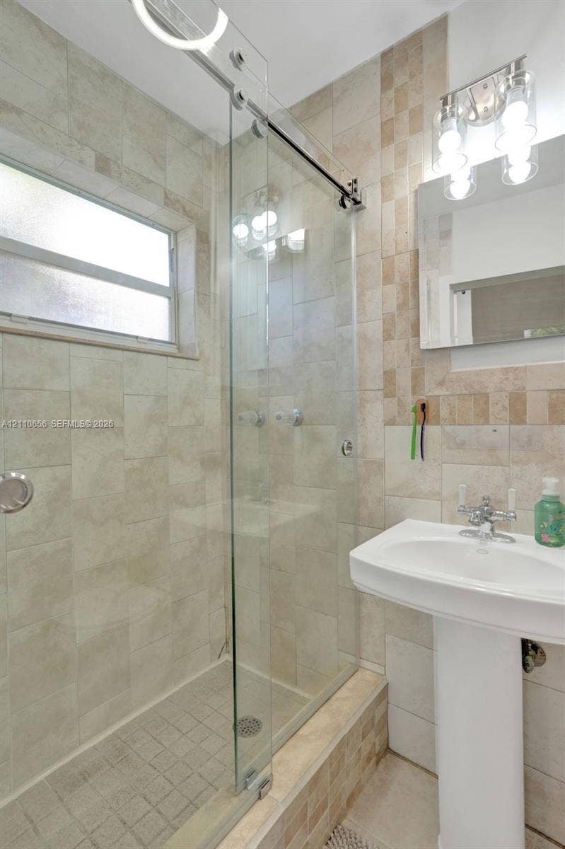 30 NE 48th Court, Oakland Park, FL 33334 Glass-enclosed shower with a sliding door, pedestal sink, wall-mounted rectangular mirror, and neutral-toned wall tile