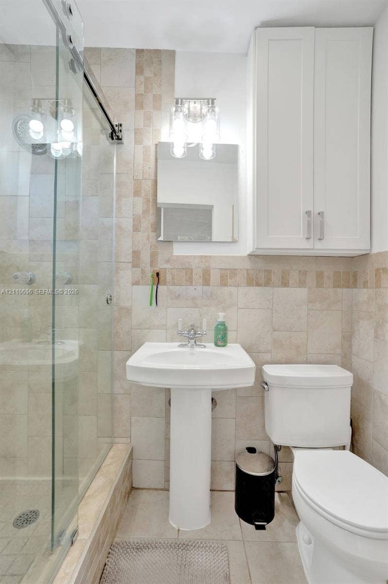 30 NE 48th Court, Oakland Park, FL 33334 Bathroom featuring neutral stone-finish wall tile with mosaic accents, a glass-enclosed shower, a white pedestal sink, and wall-mounted white cabinetry