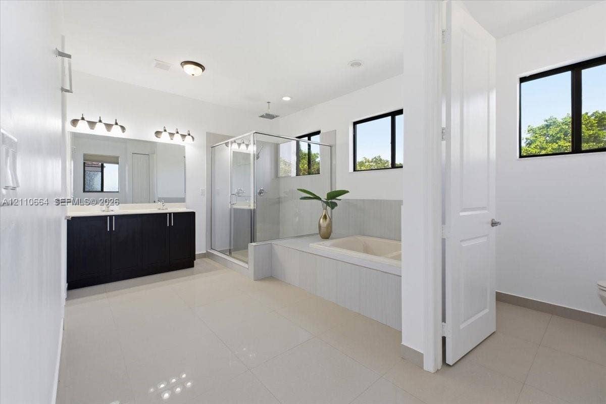 Primary bathroom featuring a dual-sink vanity with rich, dark cabinetry, a glass-enclosed shower, deep soaking tub, tile flooring, and a private water closet