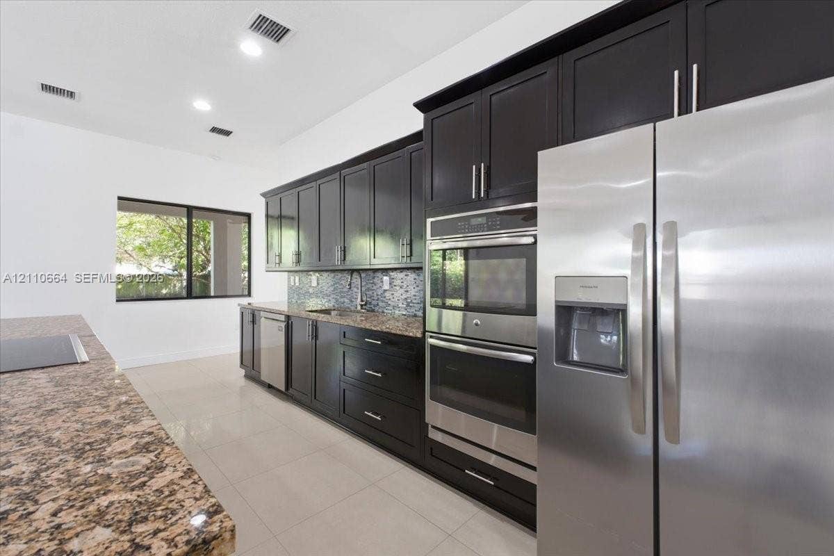 Spacious kitchen with a generous center island housing a new induction cooktop, granite countertops, rich dark cabinetry, double wall ovens, stainless steel refrigerator and dishwasher, recessed lighting, and tile flooring