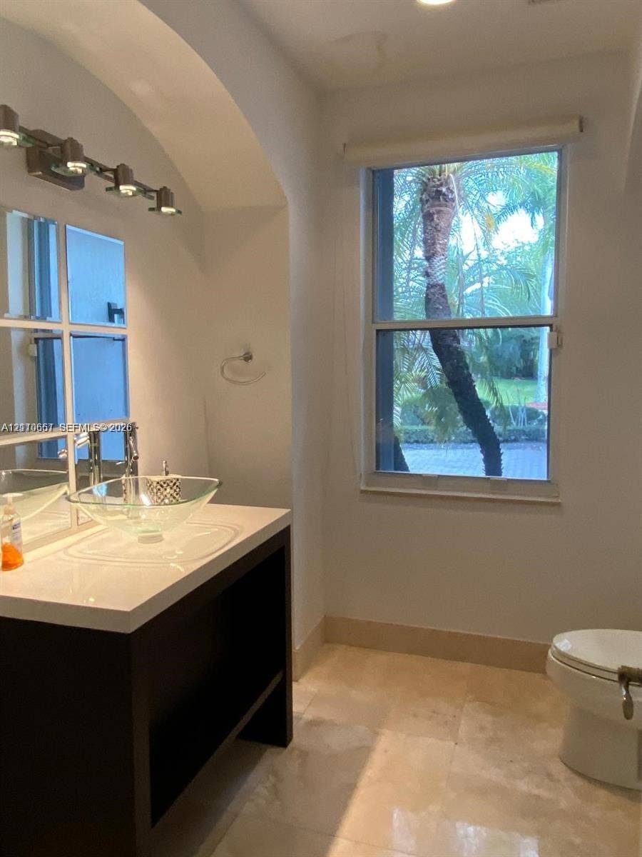Restroom featuring neutral stone-finish flooring, a wall-mounted glass shelf, a dark-framed rectangular mirror, and an arched ceiling detail