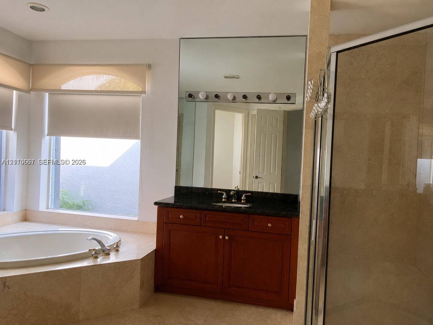 Corner shower enclosure with a metallic frame, neutral stone-finish wall tile, vanity with dark stone countertop, integrated mirror, and patterned tile flooring
