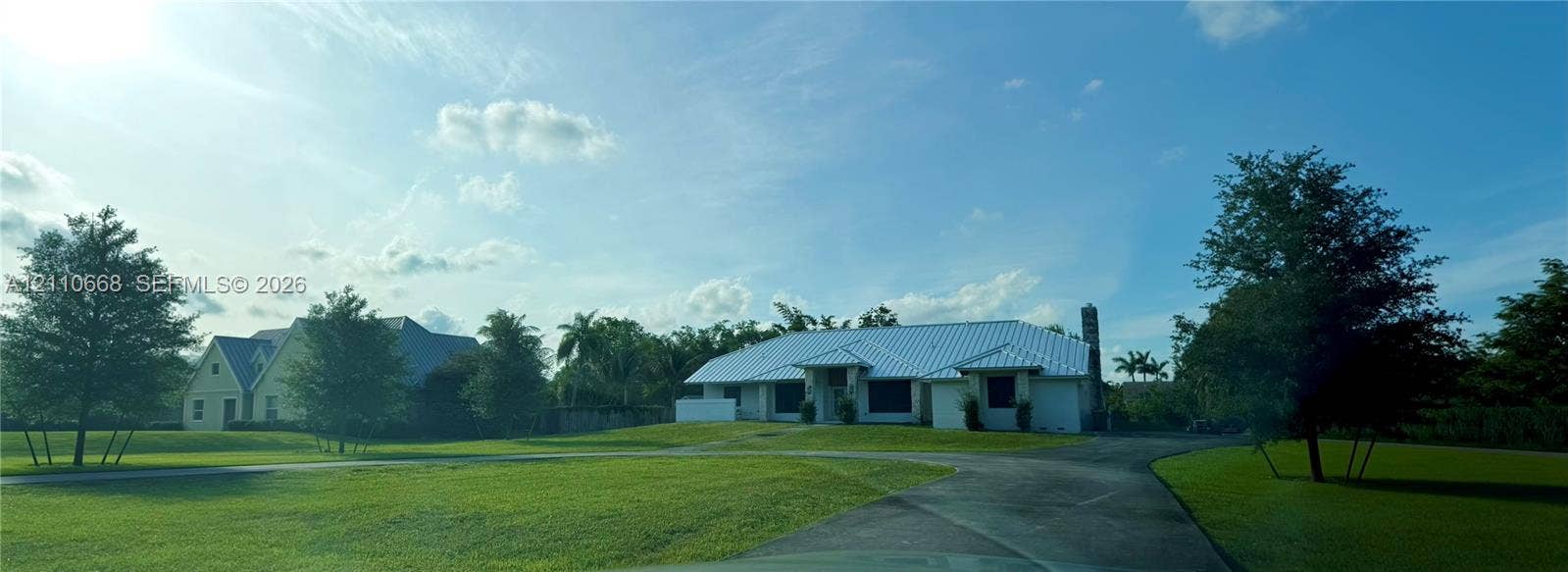 Single-story residence featuring a light-colored metal roof, expansive manicured lawn, paved driveway, and mature landscaping