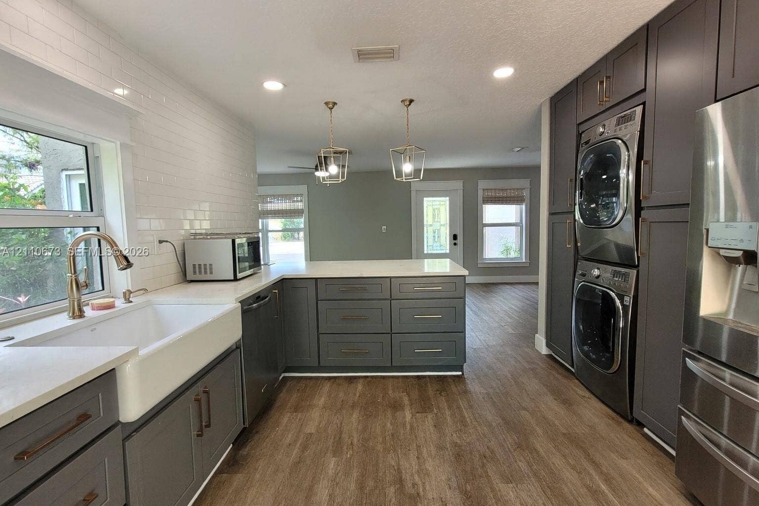 Kitchen featuring dark gray cabinetry, white subway tile backsplash, farmhouse-style sink, quartz countertops, and built-in stacked laundry appliances