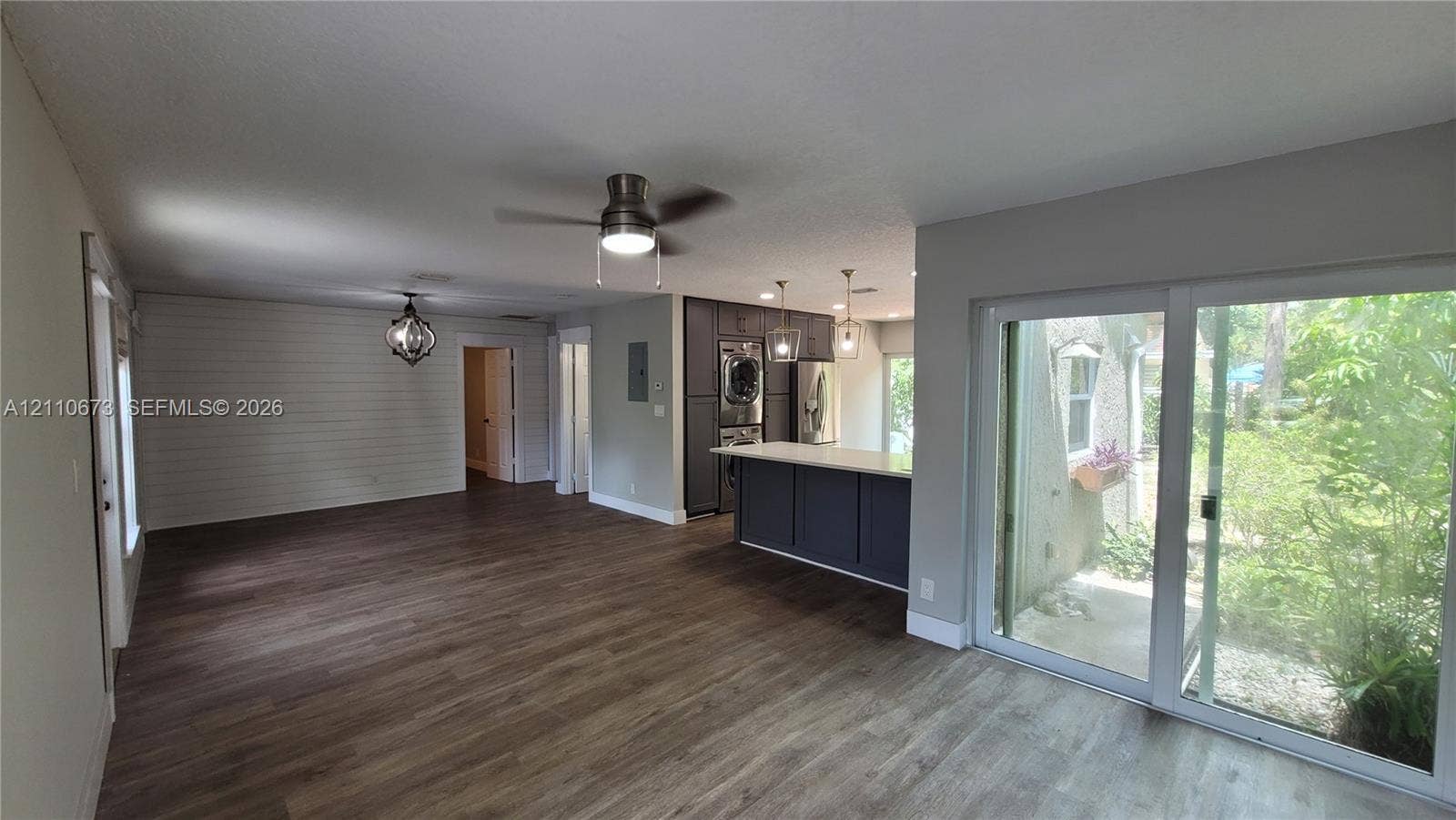 Open-concept interior featuring wood-finish flooring, a shiplap accent wall, kitchen island with quartz-style countertop, ceiling fan with integrated lighting, and sliding glass doors