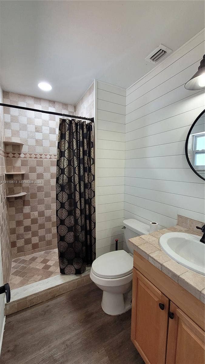 Bathroom featuring white shiplap walls, wood-finish flooring, a tiled vanity countertop with an integrated basin, and a walk-in shower with built-in shelving and multi-toned wall tile