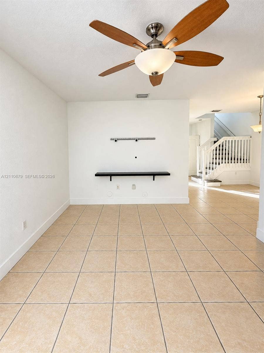 Ceramic tile flooring, ceiling fan with integrated lighting, wall-mounted display shelving, and a painted wooden staircase with white balusters
