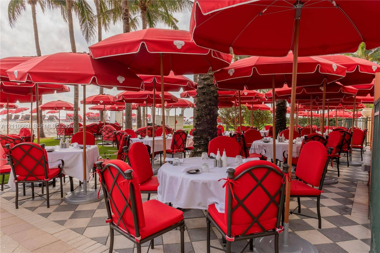 Outdoor patio featuring a checkered tile floor, numerous red umbrellas, and mature palm trees