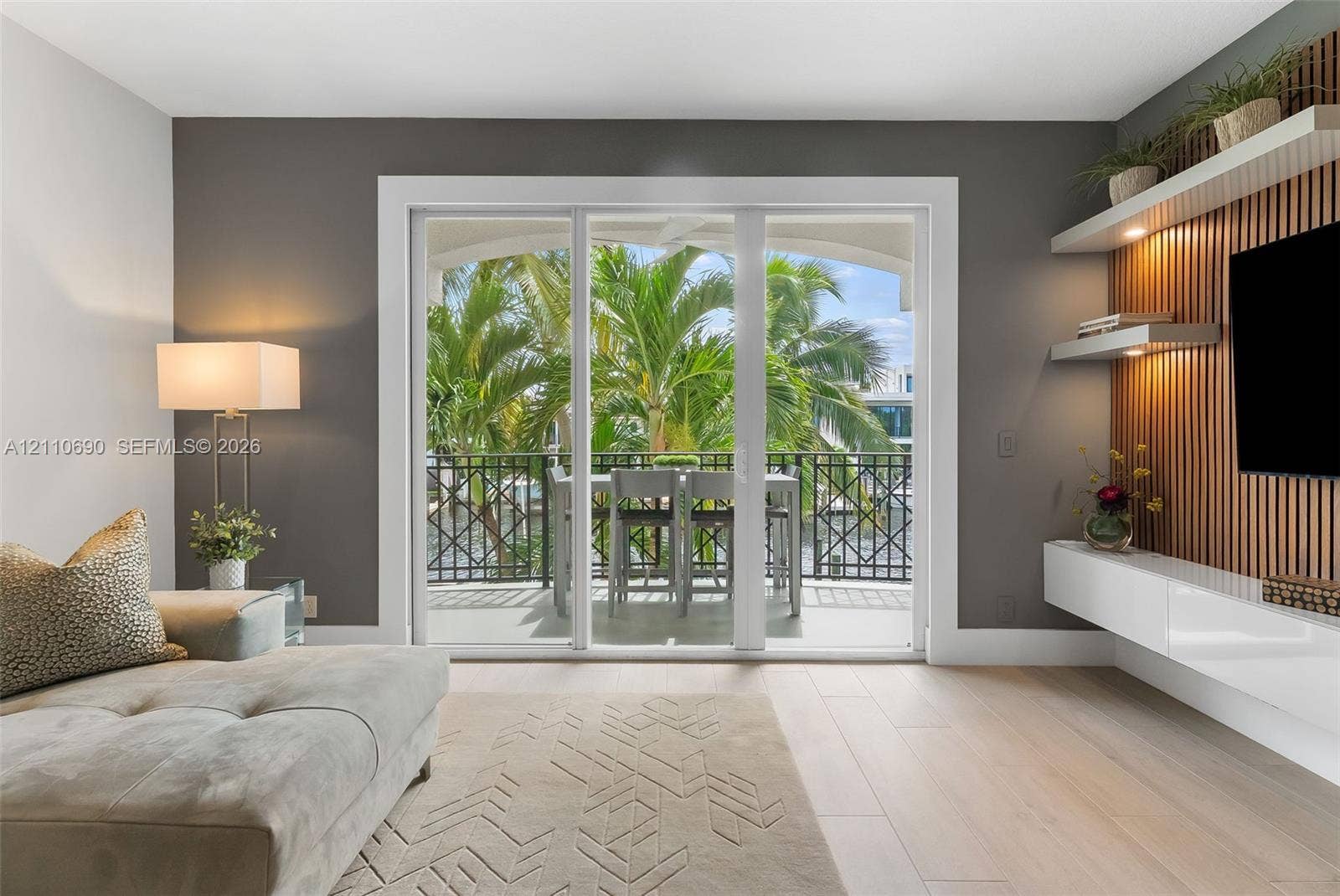 Sliding glass doors opening to a covered balcony with decorative metal railing, wood-slat accent wall with integrated shelving, and white high-gloss floating cabinetry