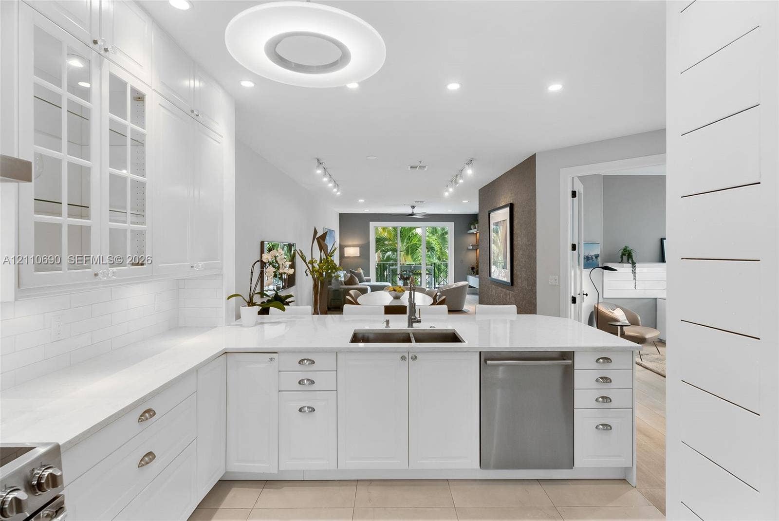 White cabinetry featuring glass-paneled upper doors, expansive countertops with an integrated stainless steel dual-basin sink, stainless steel dishwasher, tile flooring, and decorative horizontal wall paneling