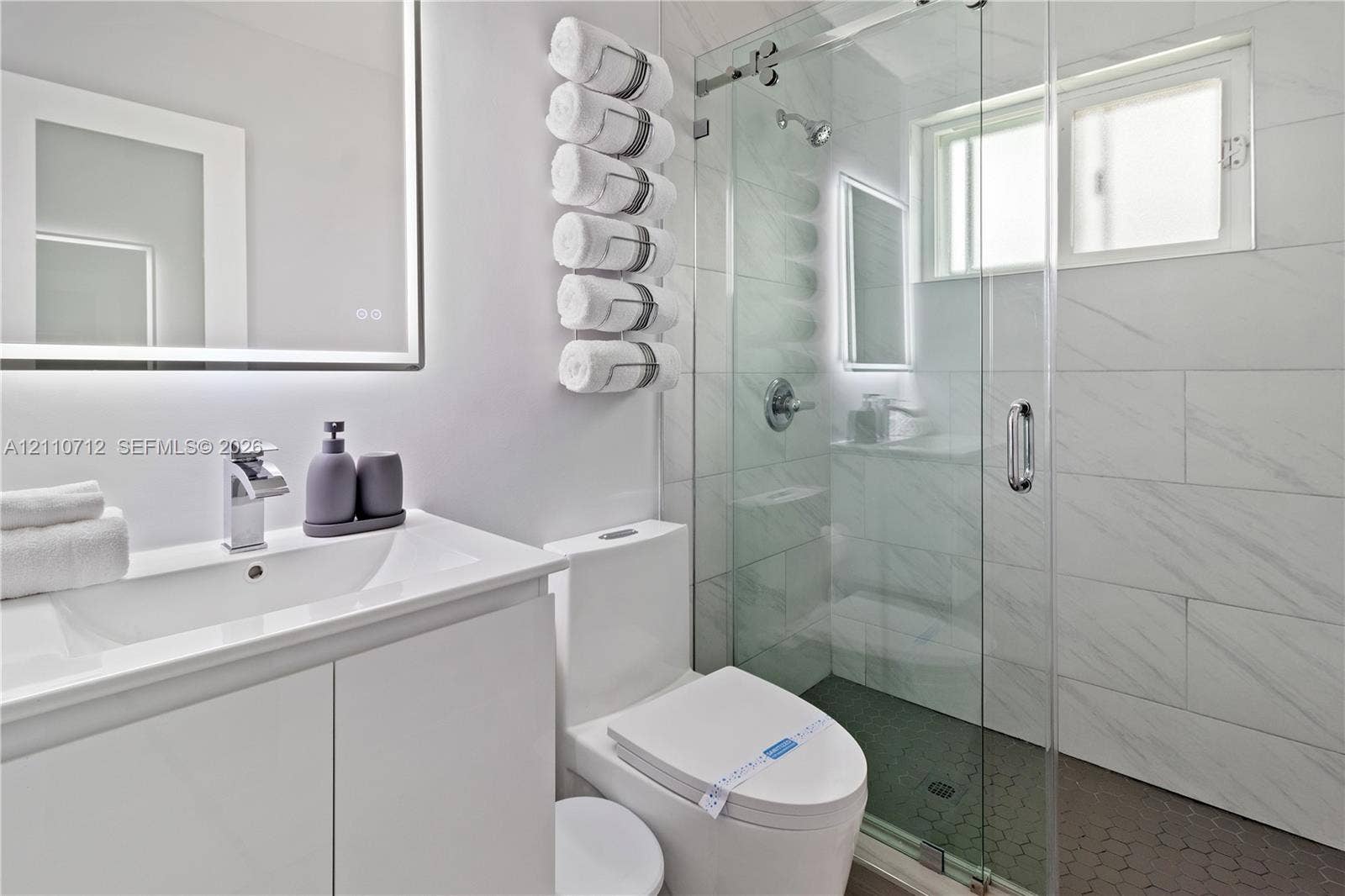 Bathroom featuring a white vanity with integrated basin, LED-backlit mirror, frameless glass shower enclosure, large-format tiled shower walls, and hexagonal floor tiling