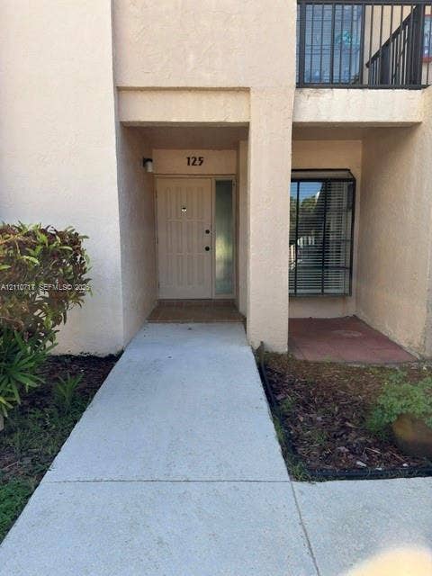 2297 SW 15th Street, #125, Deerfield Beach, Deerfield Beach, FL 33442 Exterior entryway featuring a concrete walkway, stucco facade, paneled door with a sidelight, and an upper-level balcony with metal railing