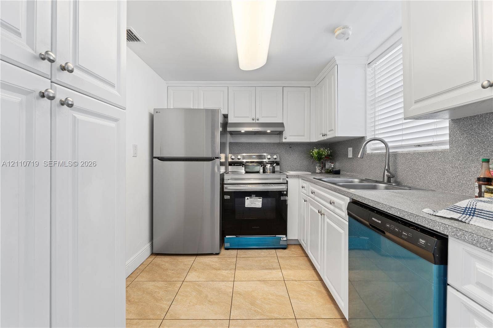 9375 Fontainebleau Boulevard, #L314, Miami, FL 33172 Kitchen featuring white shaker-style cabinetry, stainless steel appliances, neutral stone-finish countertops, dual-basin stainless steel sink with high-arc faucet, and tiled flooring
