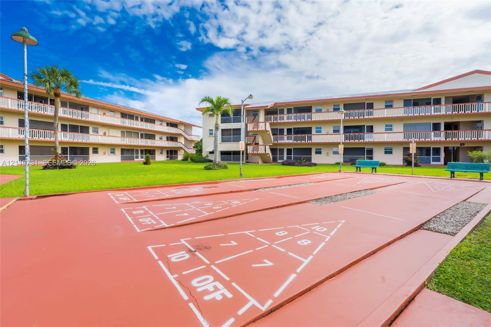 Outdoor shuffleboard court featuring a paved surface, multi-story buildings with external balconies, and landscaped lawn areas
