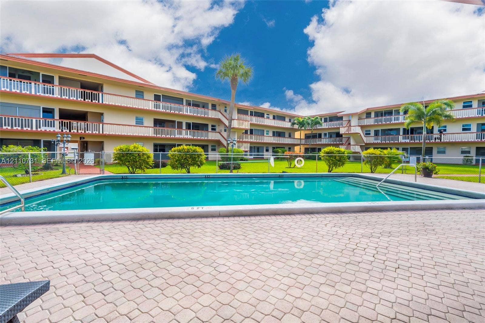 Community pool featuring a concrete deck, paved surround, metal perimeter fencing, and mature landscaping