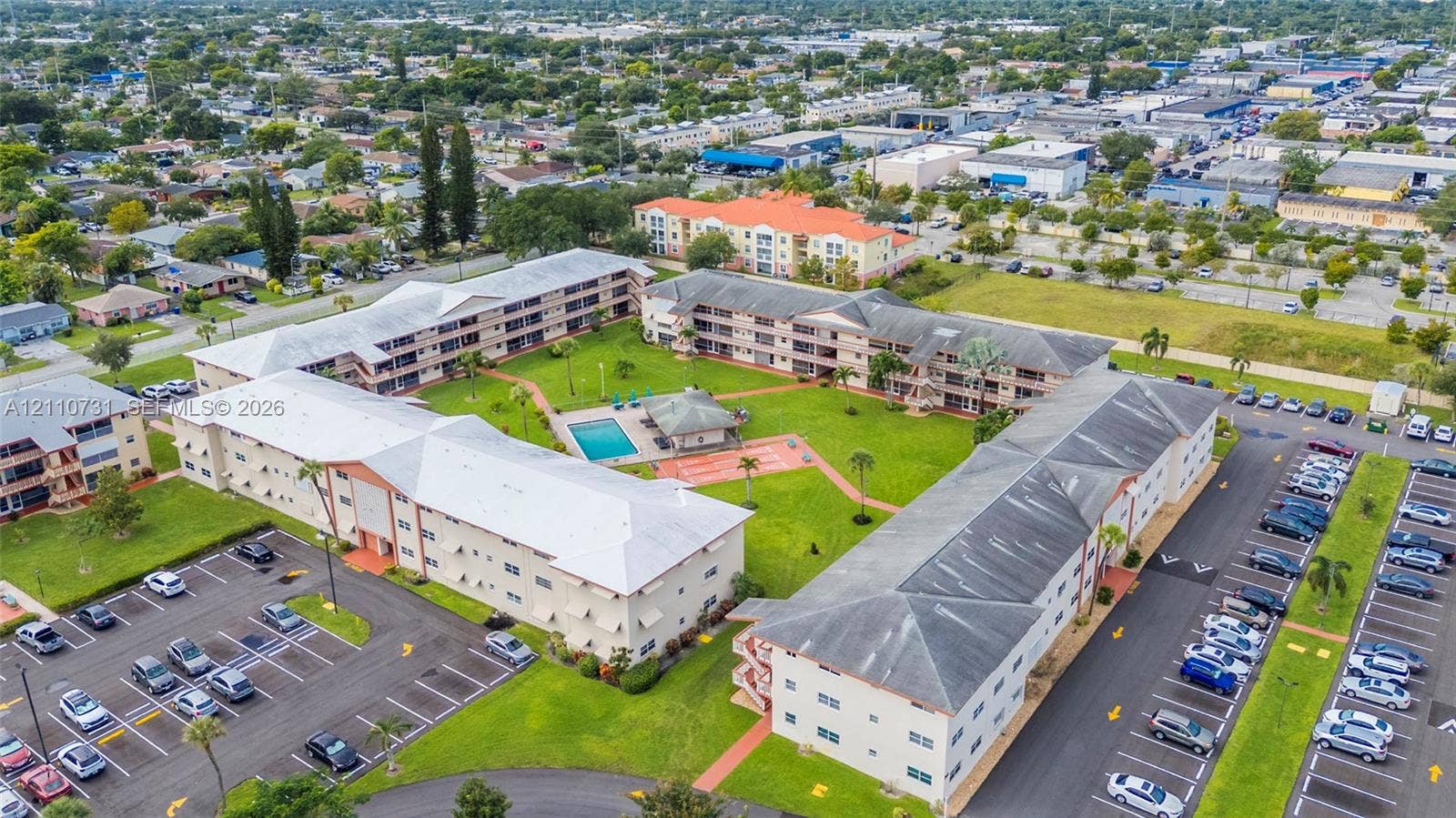 Multi-building residential complex featuring a central swimming pool, covered pavilion, paved parking areas, and mature landscaping