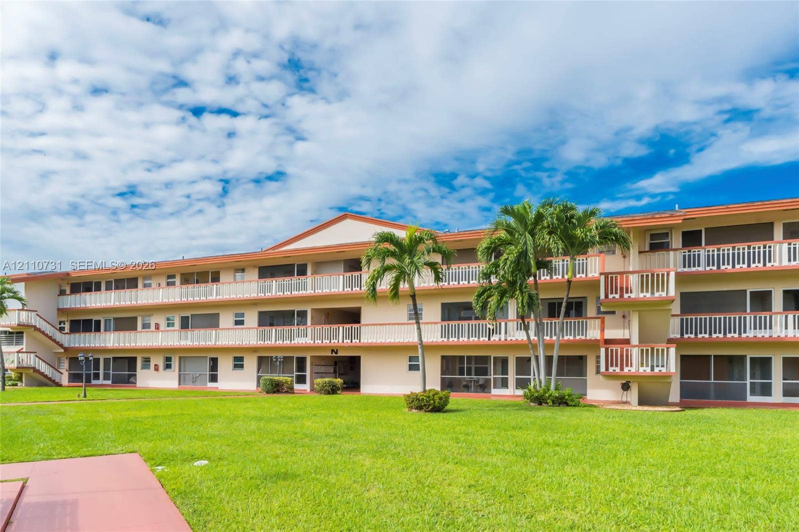 Multi-story exterior featuring cream-colored stucco facade, horizontal balustraded balconies, and a pitched roofline, set against a manicured lawn with mature palm trees