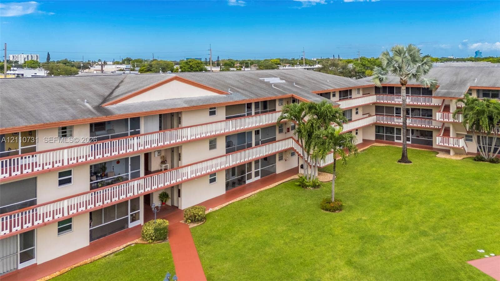 Multi-story building featuring screened balconies, shingled roofing, manicured lawn, and mature palm trees