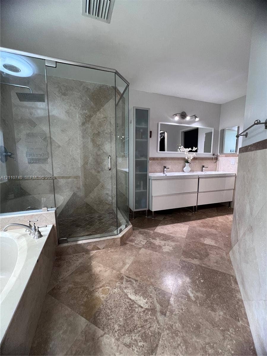 Corner glass-enclosed shower featuring stone-tile surround, dual-sink vanity with built-in cabinetry, soaking tub, and stone-tile flooring