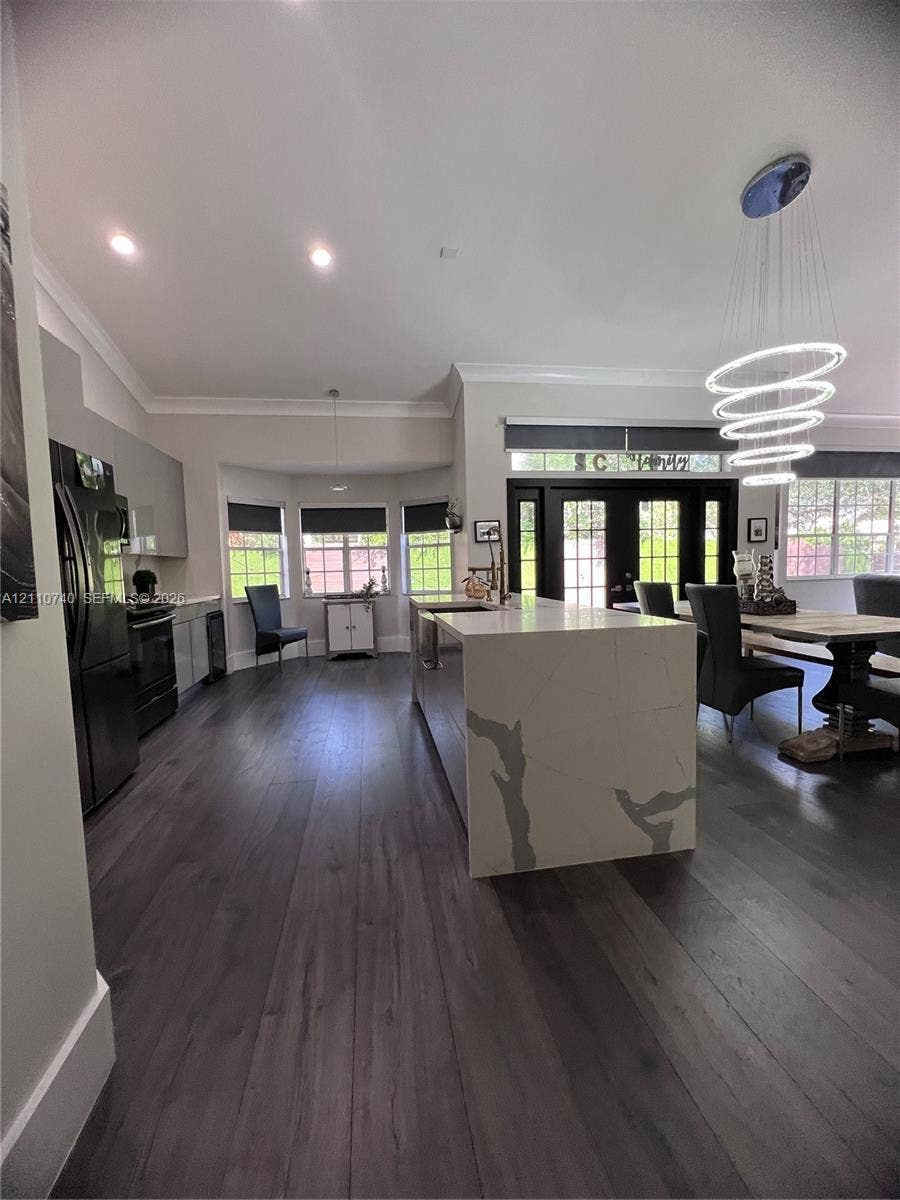 Kitchen featuring an expansive waterfall-edge island with stone-finish countertop, wood-finish flooring, recessed lighting, multi-tiered ring pendant light, and black double-entry doors with transom windows